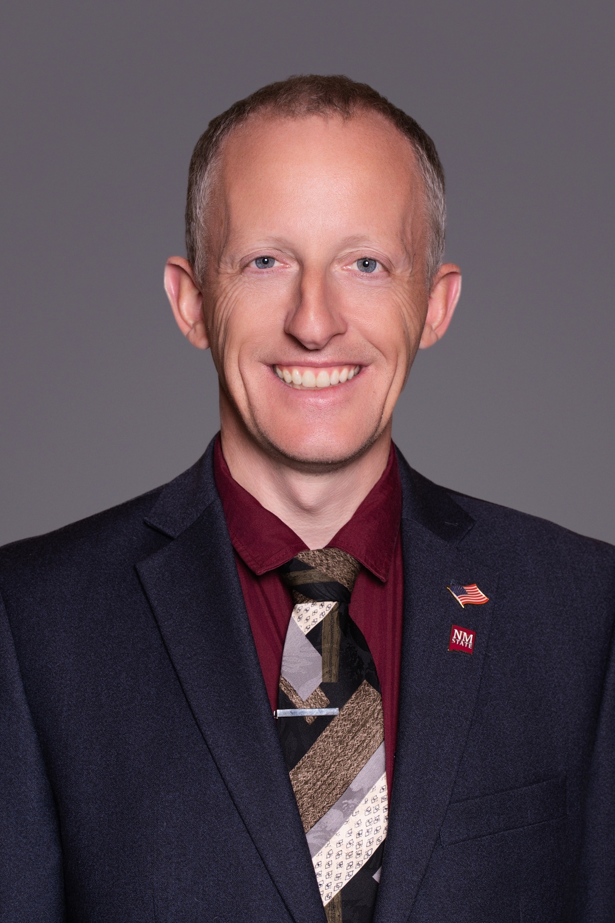 Smiling man in a suit and tie posing for a professional headshot against a gray background, with U.S. flag and NM State lapel pins.