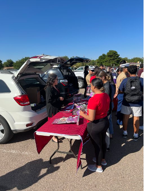 Roswell High School students stand in front the NMSU table at the Roswell College Fair. The NMSU table is placed in front of a white SUV type car with an NMSU employee standing between the car and the table. 
