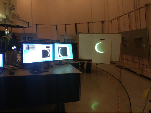Sunspot telescope displays the solar eclipse on two computer monitor screens on the left side of the photo, as well as projected on a white sheet of paper taped to a whtie board on the right side of the photo. 