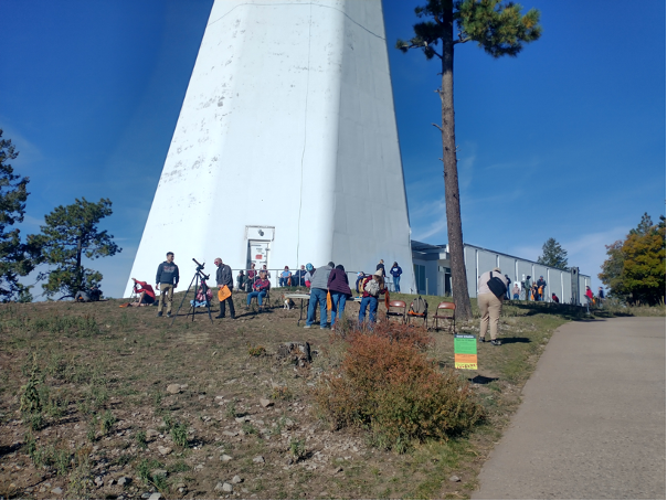 Multiple individuals stand outside of the Sunspot telescope looking at the solar eclipse through smaller telescopes. The white base of the Sunspot Telescope can be seen in the background. 