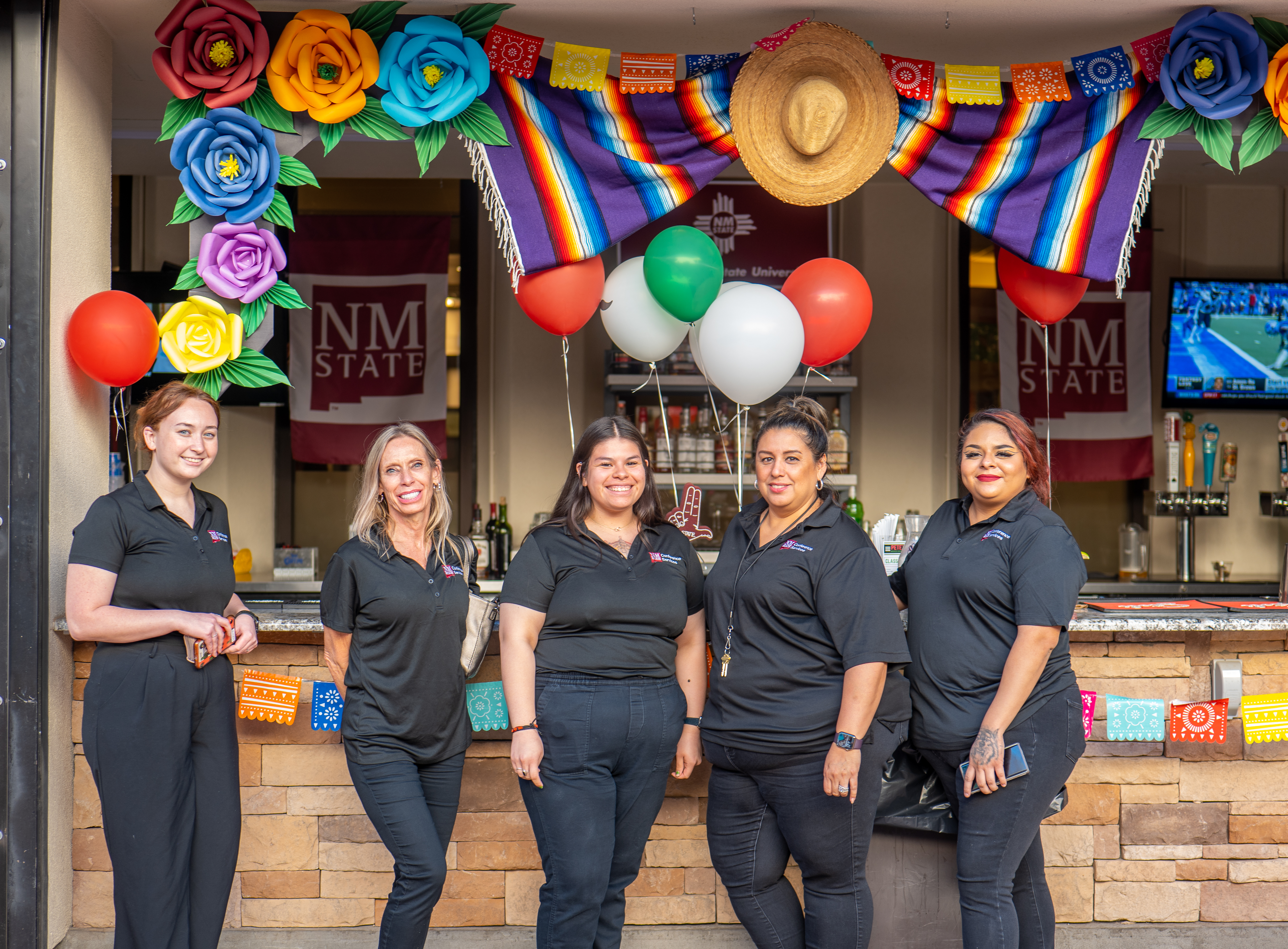 NMSU Conference Services staff stand in a line in all black in front of the bar at Pete's Patio. Above them are traditional flowers, banners, and Mexican blankets to decorate the bar for Hispanic Heritgate Month festivities. 