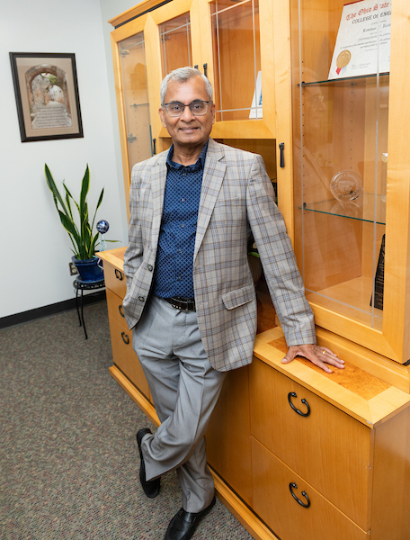 Man in a plaid blazer and glasses leaning against a wooden display cabinet in an office.