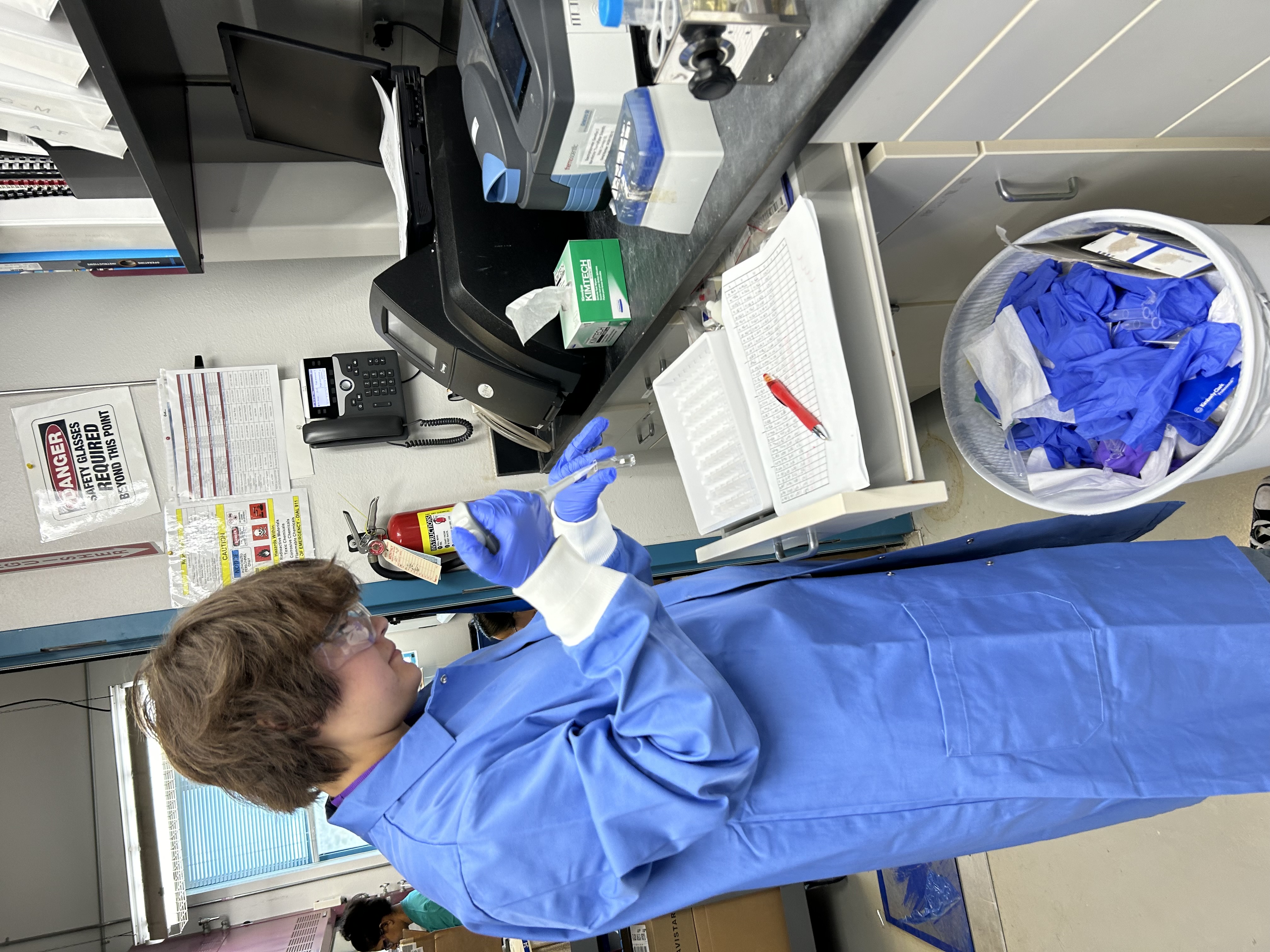 Emmie Navarrette in a blue lab coat and goggles is preparing samples in a laboratory, with a tray of test tubes and a waste bin for used gloves in the foreground.