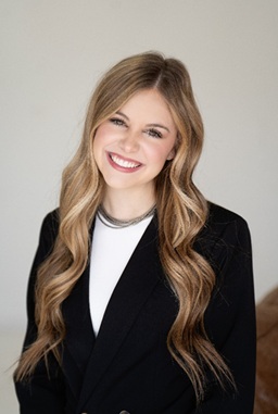 A smiling young woman with long, wavy hair wearing a black blazer and a white top.