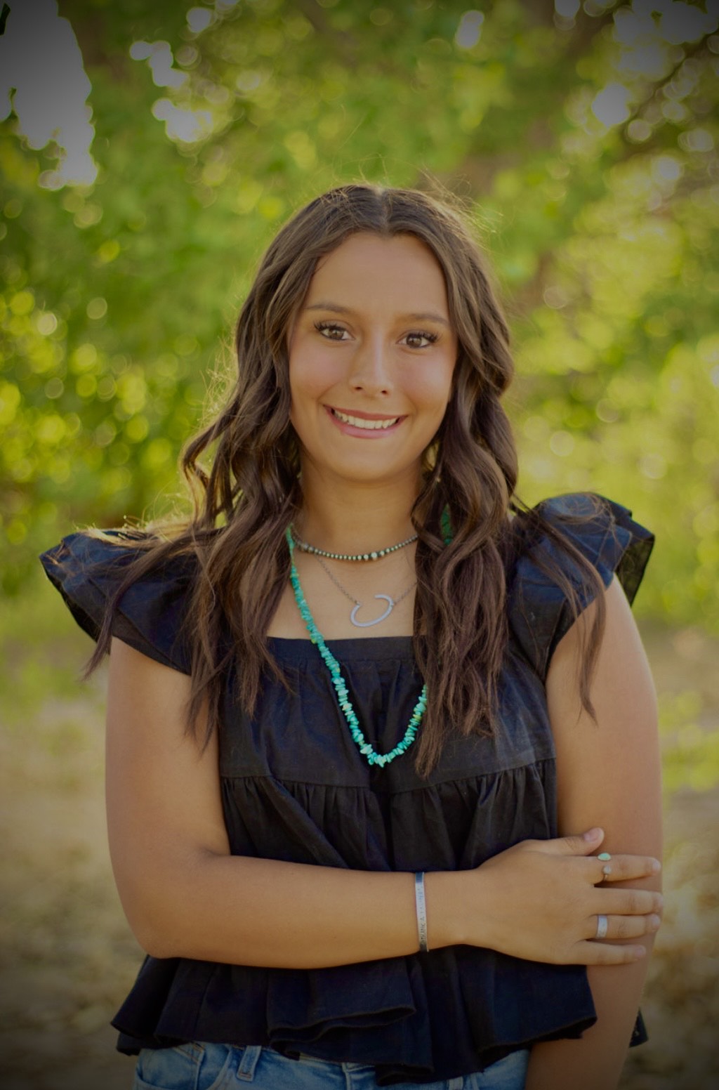 A smiling young woman in a black ruffled top stands outdoors, wearing a turquoise necklace and showcasing a natural background.