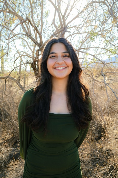 A young woman smiling in a green dress stands outdoors with a blurred tree background.