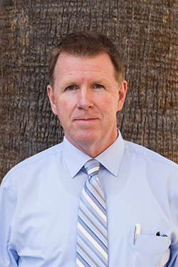 Professional headshot of a man wearing a light blue dress shirt and striped tie, standing outdoors against a textured tree background.