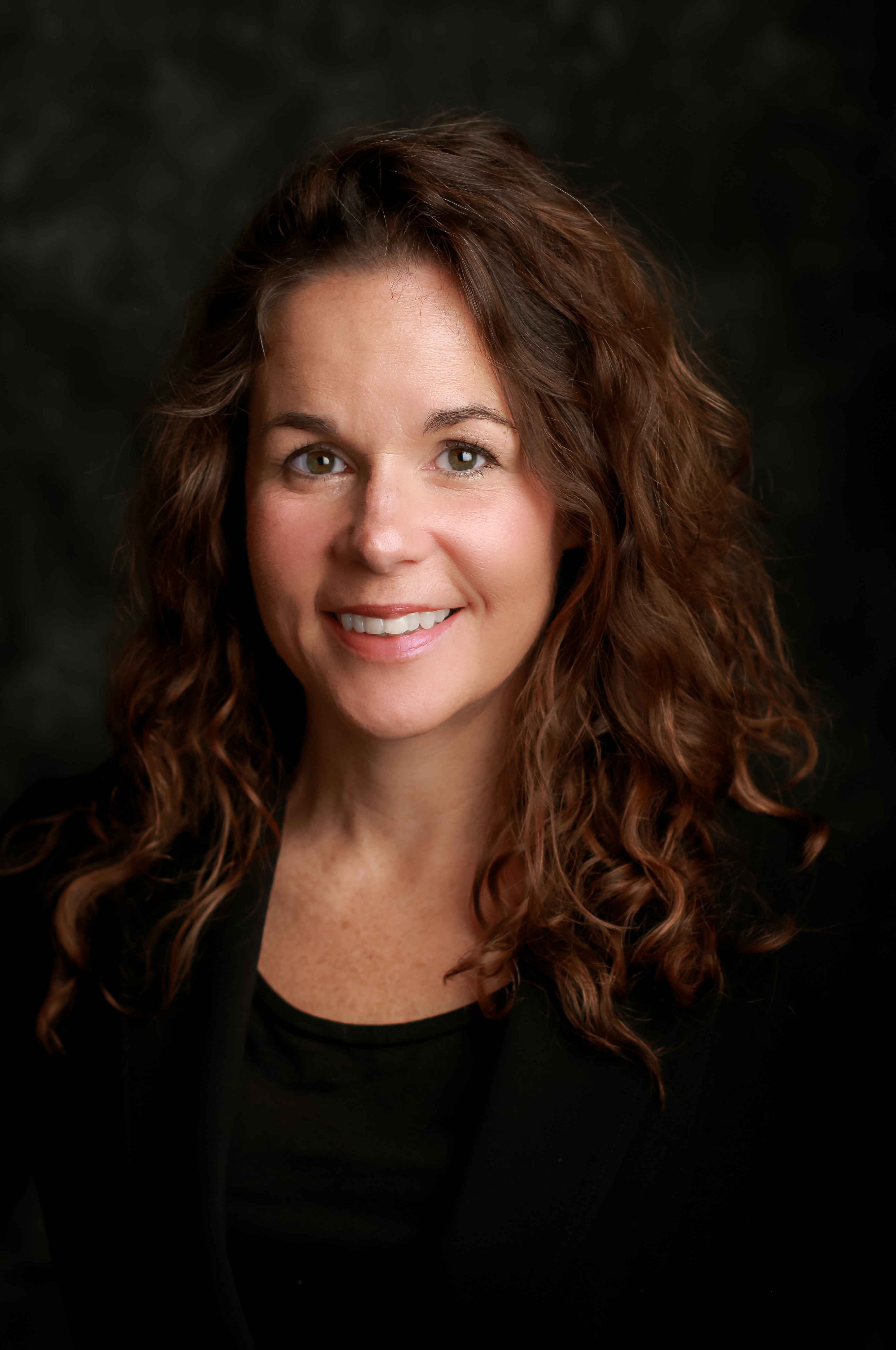 Professional headshot of a smiling woman with long curly brown hair against a dark background.