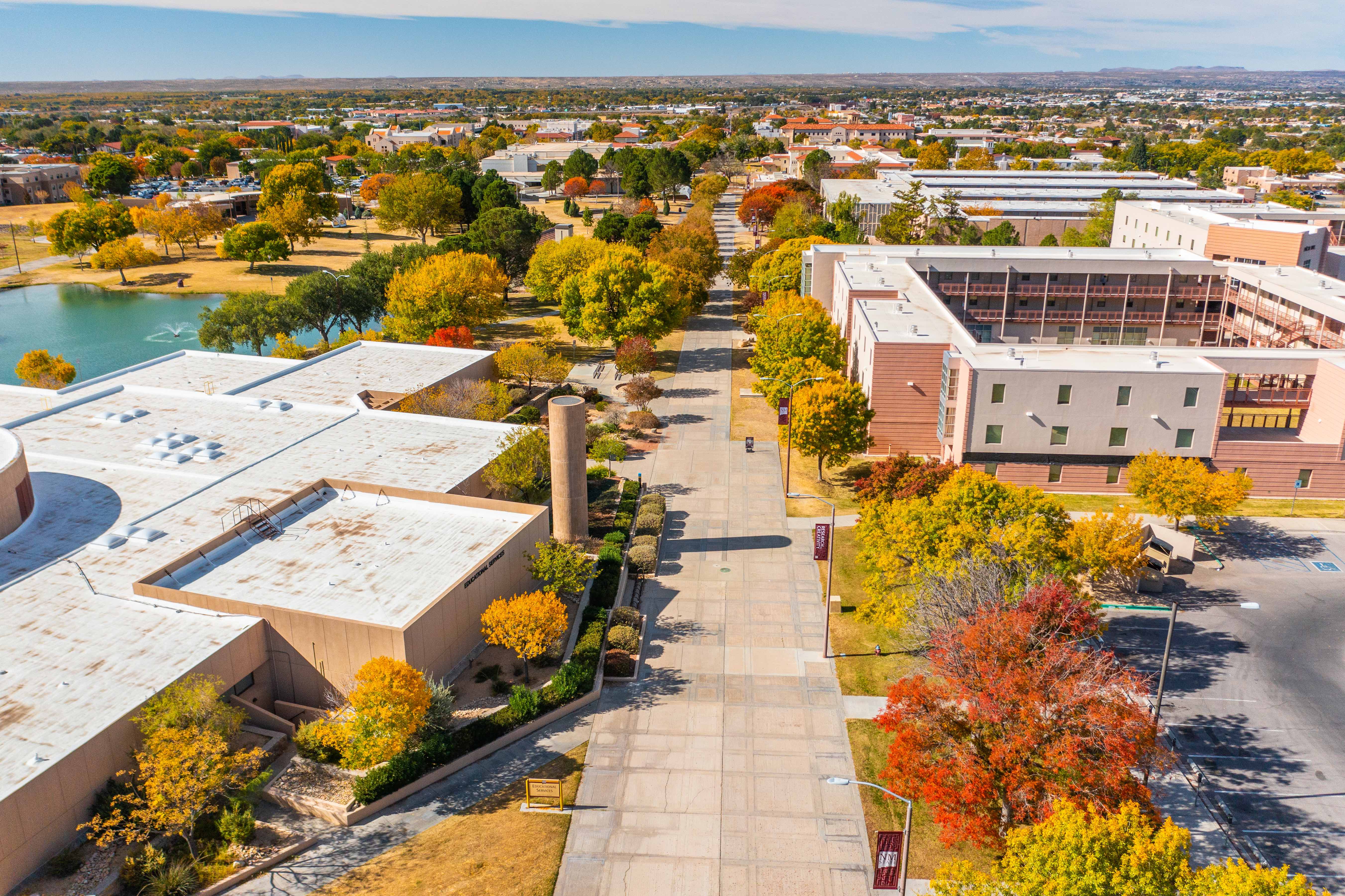 Aerial view of a campus landscape featuring colorful autumn trees, buildings, and a pathway beside a pond.