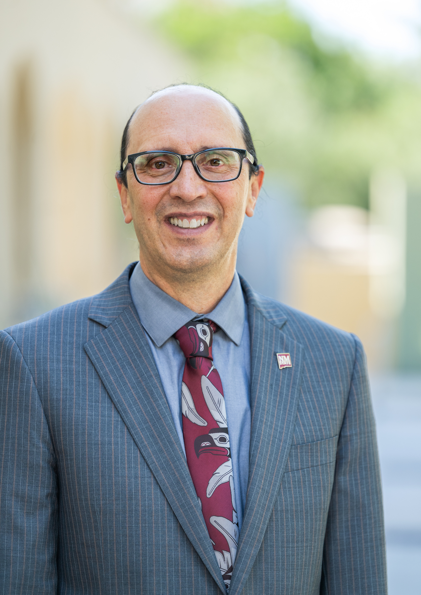 Smiling man in a suit and tie posing for a professional headshot against a gray background, with U.S. flag and NM State lapel pins.